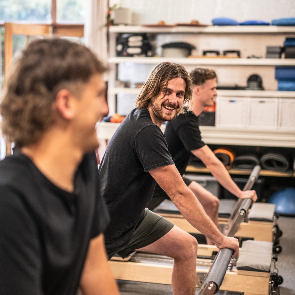 Smiling young man leans on Pilates reformer, smiling at camera